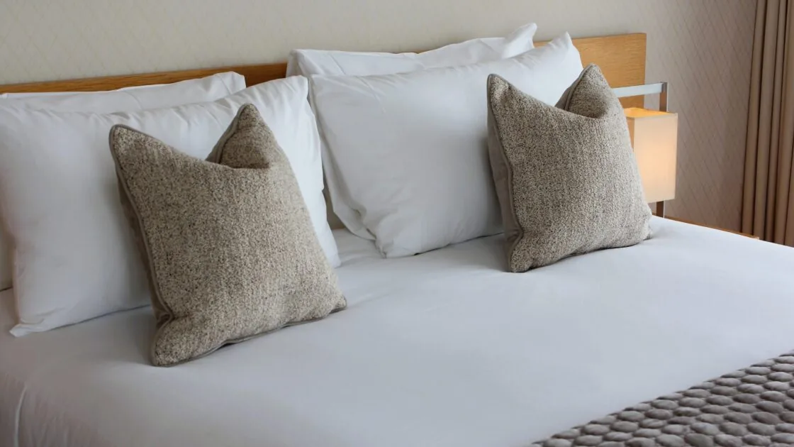 A neatly made bed with white sheets, two large square gray pillows, and a textured gray blanket at the foot of the bed.