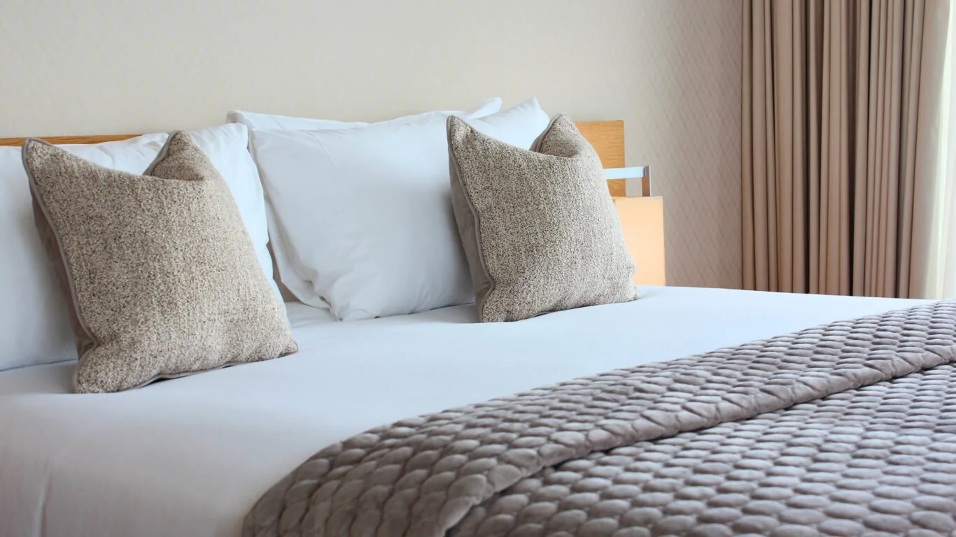 A neatly made bed with white sheets, two large beige pillows, and a textured gray blanket, set beside a window with beige curtains.