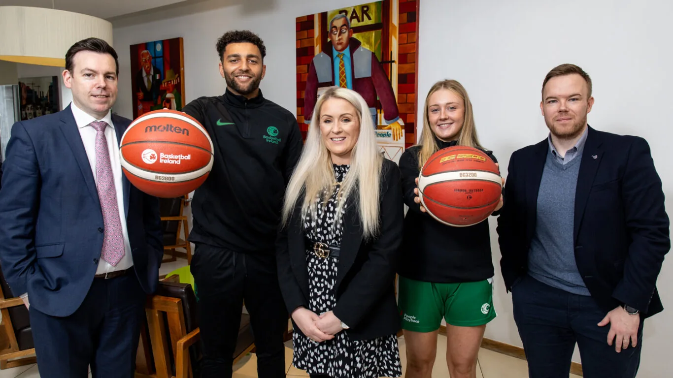 Five people pose indoors; two are holding basketballs and wearing sportswear, while the others are dressed in business attire. A colorful painting is visible on the wall behind them.