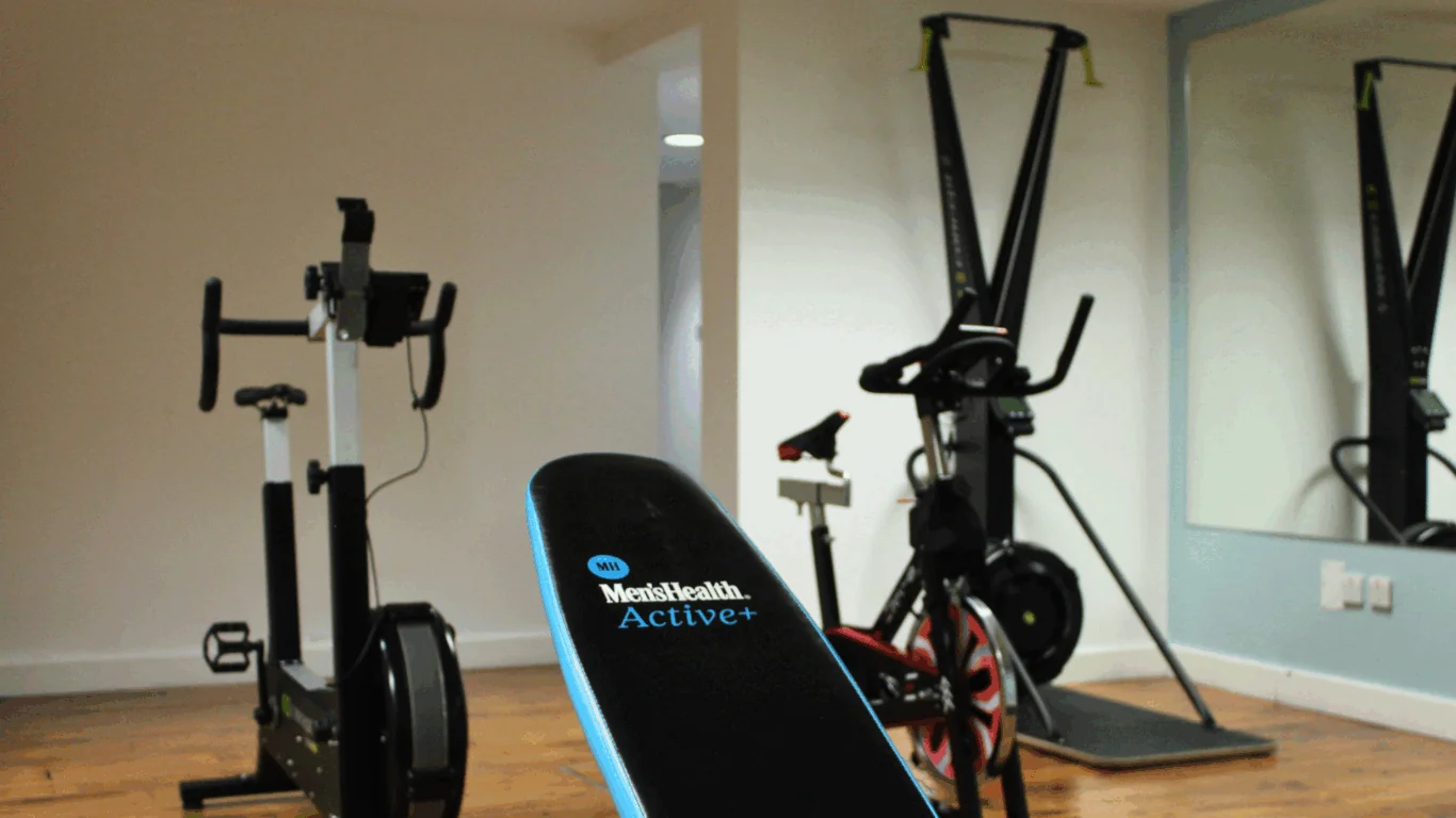 A small gym room with exercise equipment, including a rowing machine, stationary bike, ski machine, and a bench labeled 