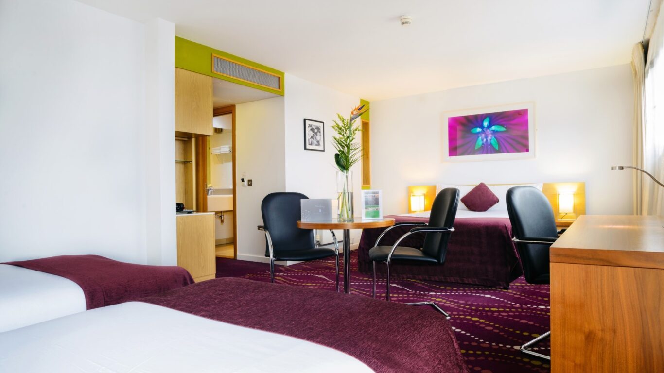 Hotel room with two beds, a small table with chairs, lamps, a flower vase, a dresser, and a colorful artwork on the wall above the bed. Decor features purple and white tones.