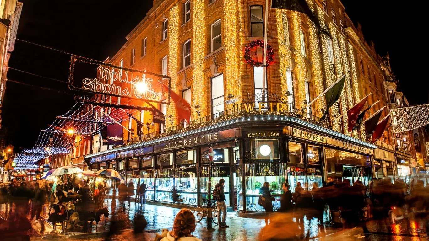 A busy city street at night with crowds passing a corner jewelry store, holiday lights, and festive decorations illuminating the buildings—one of the magical things to do in Dublin Christmas season.