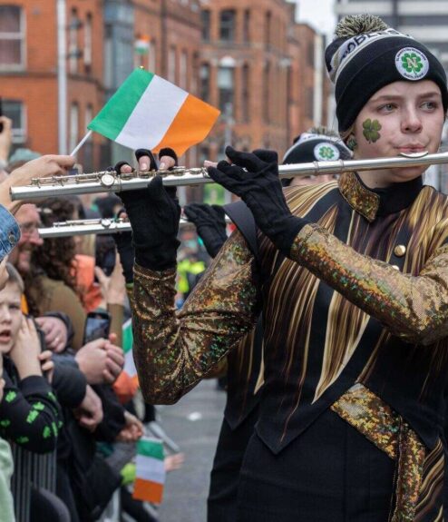 Parade musician in festive attire playing flute, holding Irish flag, surrounded by crowd.