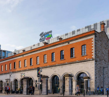 People walking by the historic Three Arena building on a sunny day in Dublin.