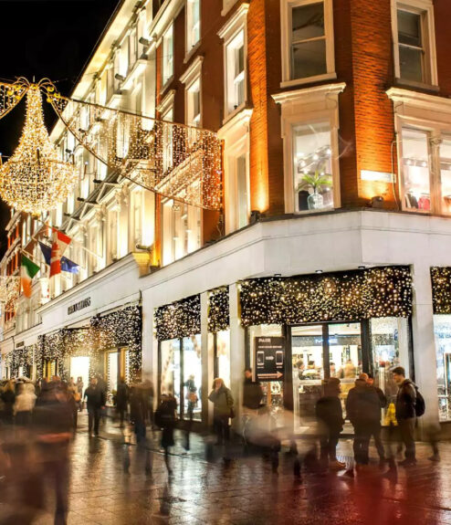 Festive shoppers stroll under twinkling lights on a vibrant Dublin street at night.