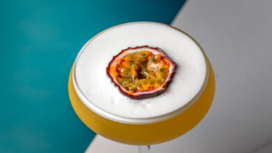 A yellow cocktail in a coupe glass with a frothy white top, garnished with a halved passion fruit, sits on a marble table next to a teal chair.
