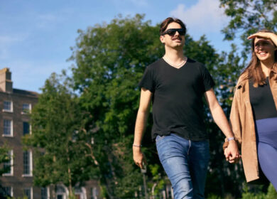 A couple walks hand in hand in a sunlit garden near a historic Dublin building.