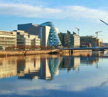 Modern buildings and Samuel Beckett Bridge reflecting on Dublin's River Liffey at sunrise.