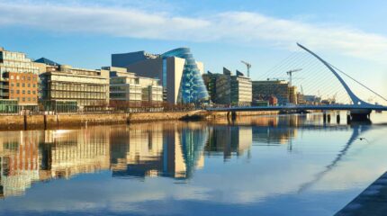 Modern buildings and Samuel Beckett Bridge reflecting on Dublin's River Liffey at sunrise.