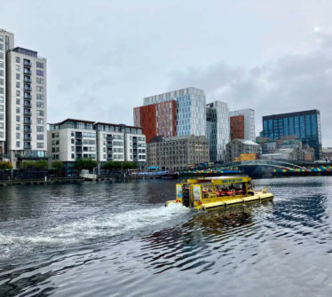 Tour boat on Dublin canal with modern buildings and colourful bridge in background.