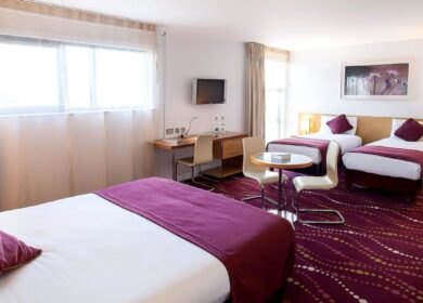Spacious hotel room with three beds, purple accents, a desk, and a cosy seating area.