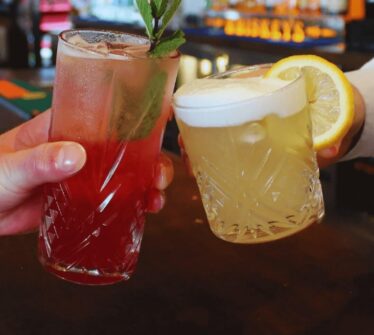 Two hands hold colourful cocktails at a lively hotel bar.