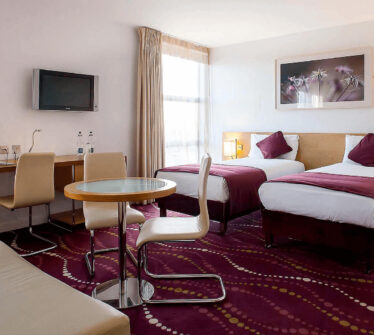 Elegant hotel room with twin beds, plush purple accents, and natural light at Louis Fitzgerald.