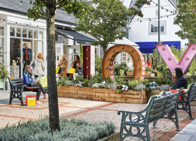 Shoppers relax near large "JOY" letters at Kildare Village, surrounded by lush greenery.