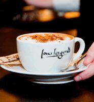 A hand serves a cappuccino in a Louis Fitzgerald cup on a saucer.