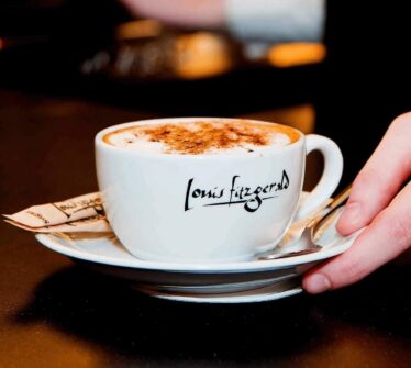 A hand serves a cappuccino in a Louis Fitzgerald cup on a saucer.