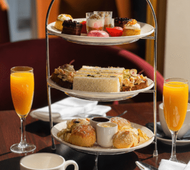 Afternoon tea spread with pastries, sandwiches, scones, and orange juice at a sunlit table.