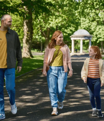 Family of five strolling in a sunny Dublin park with leafy trees and a domed pavilion ahead, parents holding hands with children, enjoying a leisurely walk