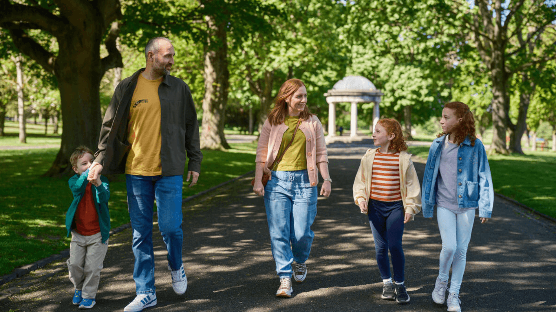 Family of five strolling in a sunny Dublin park with leafy trees and a domed pavilion ahead, parents holding hands with children, enjoying a leisurely walk